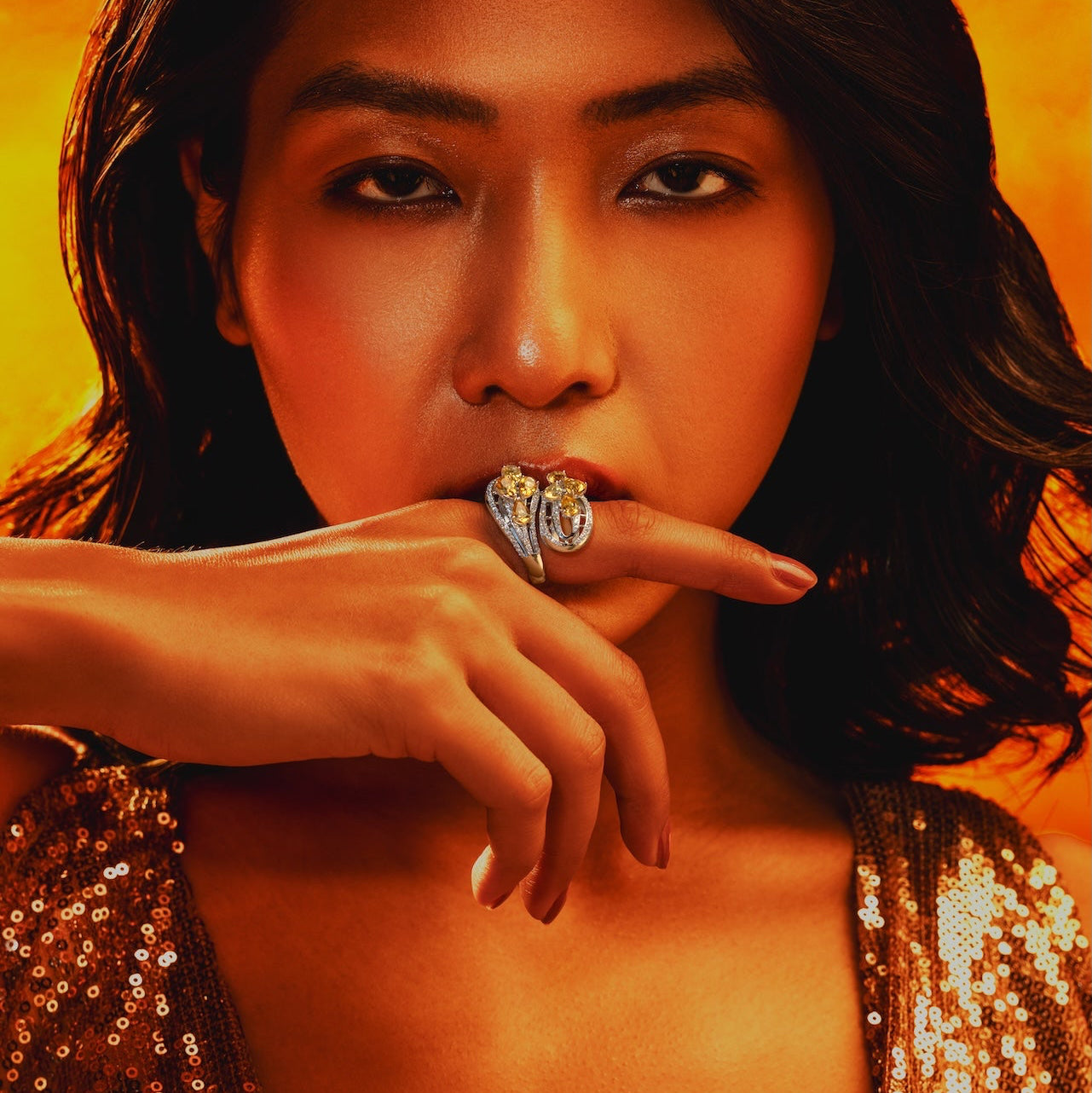 A close-up portrait of a woman with dark hair wearing a sparkling gold sequined top and a large silver ring on her finger, set against a vibrant orange background.