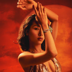 A woman in a sparkling metallic gold halter dress poses with her arms raised against a dramatic red and orange sky background.