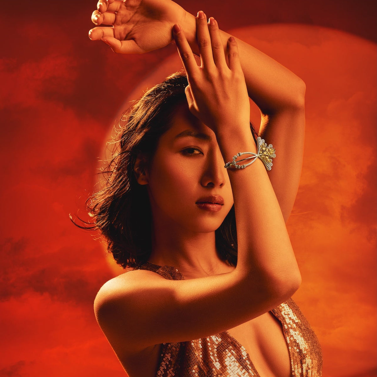 A woman in a sparkling metallic gold halter dress poses with her arms raised against a dramatic red and orange sky background.