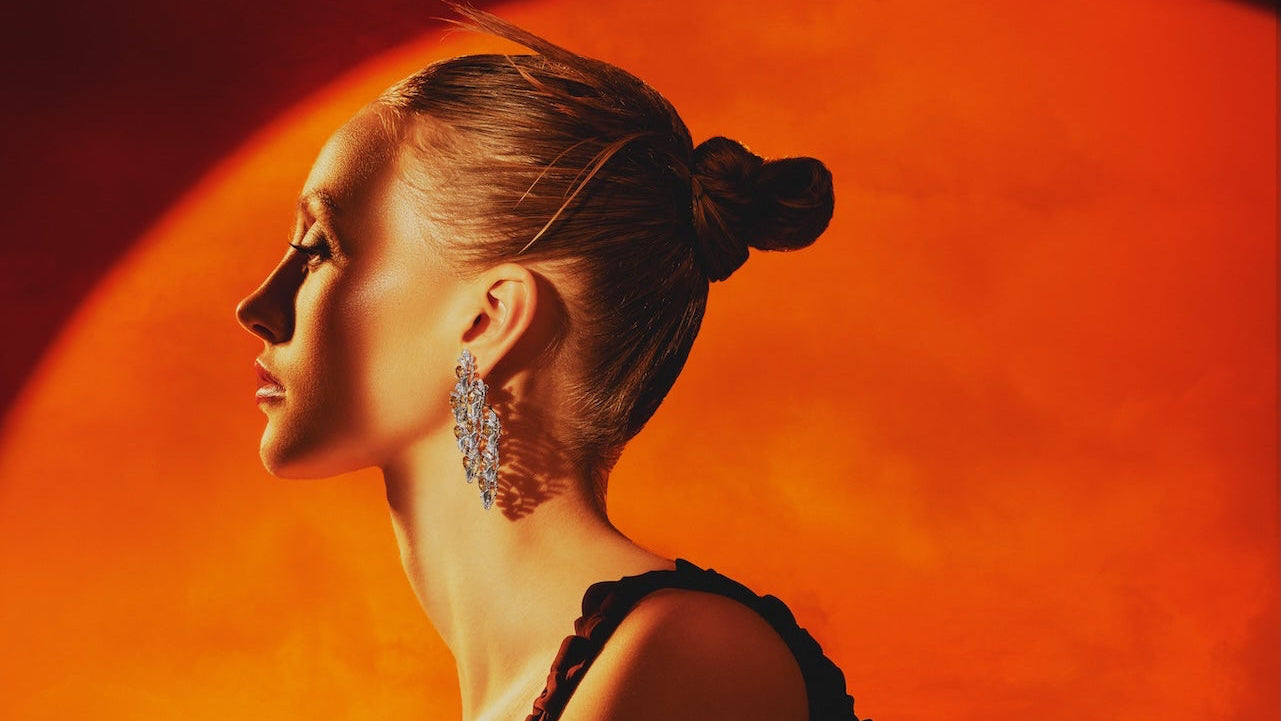 A woman in a black lace-trimmed crop top and black bottoms poses in profile against a large glowing orange circular backdrop. She is wearing large silver dangling earrings, and the scene is lit with dramatic warm orange and red lighting.