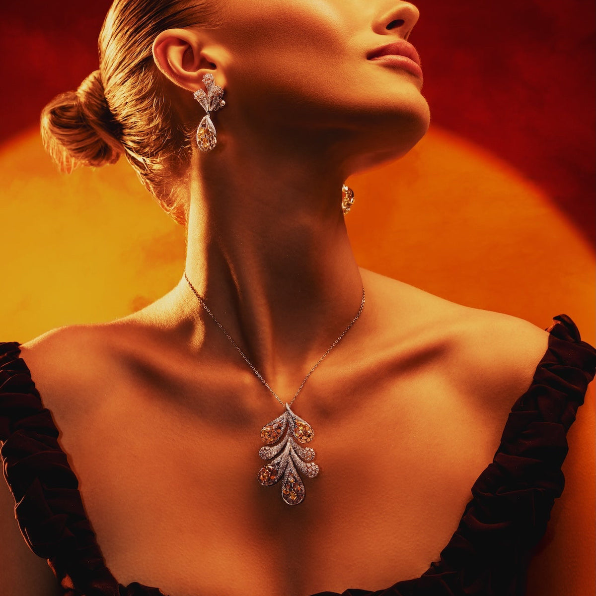 A woman with her hair in a bun wears a silver-toned leaf-shaped pendant necklace and matching drop earrings against a warm red and orange background.