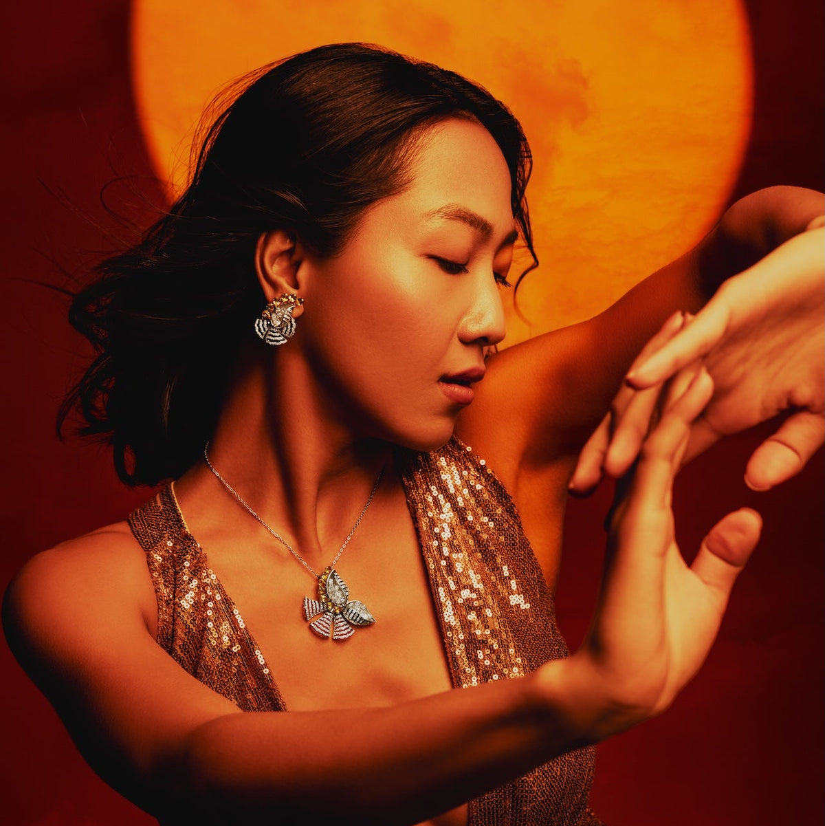 A woman in a shimmering bronze sequined dress poses with her hands in front of a large glowing orange moon against a dark red background. She is wearing a silver butterfly pendant necklace and matching butterfly earrings.