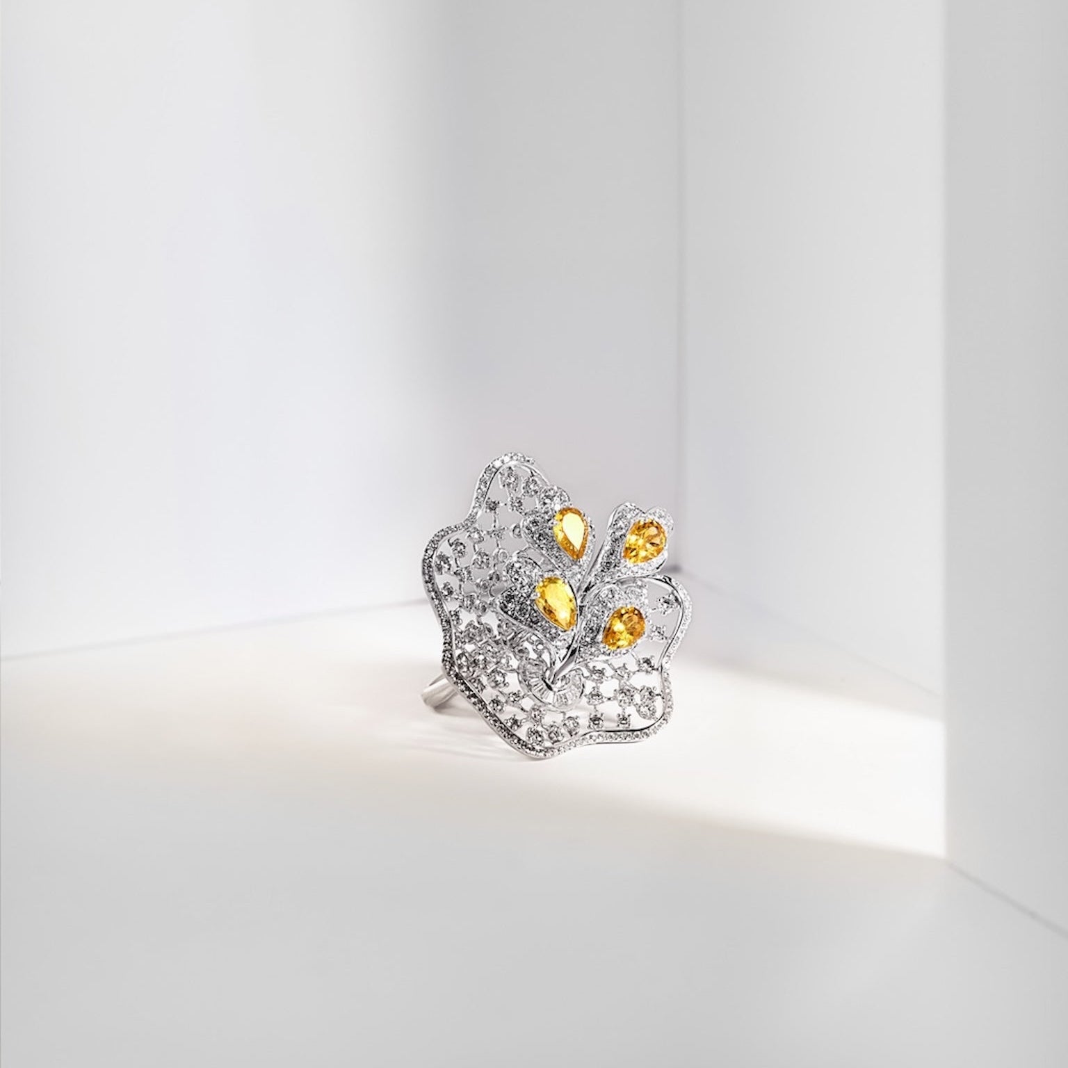 A silver-toned ring shaped like a stylized flower or crown, set with clear pavé stones and accented by four small round yellow gemstones, photographed on a clean white surface in a minimalist white corner setting.