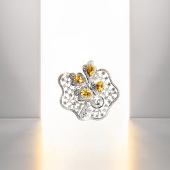 An ornate sculptural brooch featuring a silver-toned floral or cloud-like form set with numerous small clear stones and three prominent pear-cut yellow gemstones, photographed against a minimalist white studio background with a warm glow on the floor.