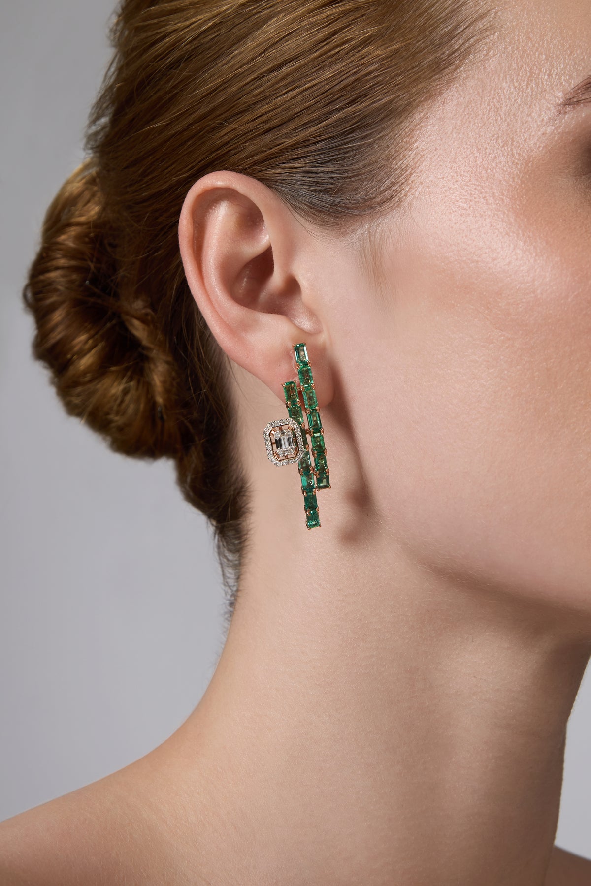 A close-up of a model's ear wearing a pair of dramatic drop earrings. The earrings feature vertical bars of emerald-cut green stones set in white metal, with a central clear gemstone surrounded by small round stones.