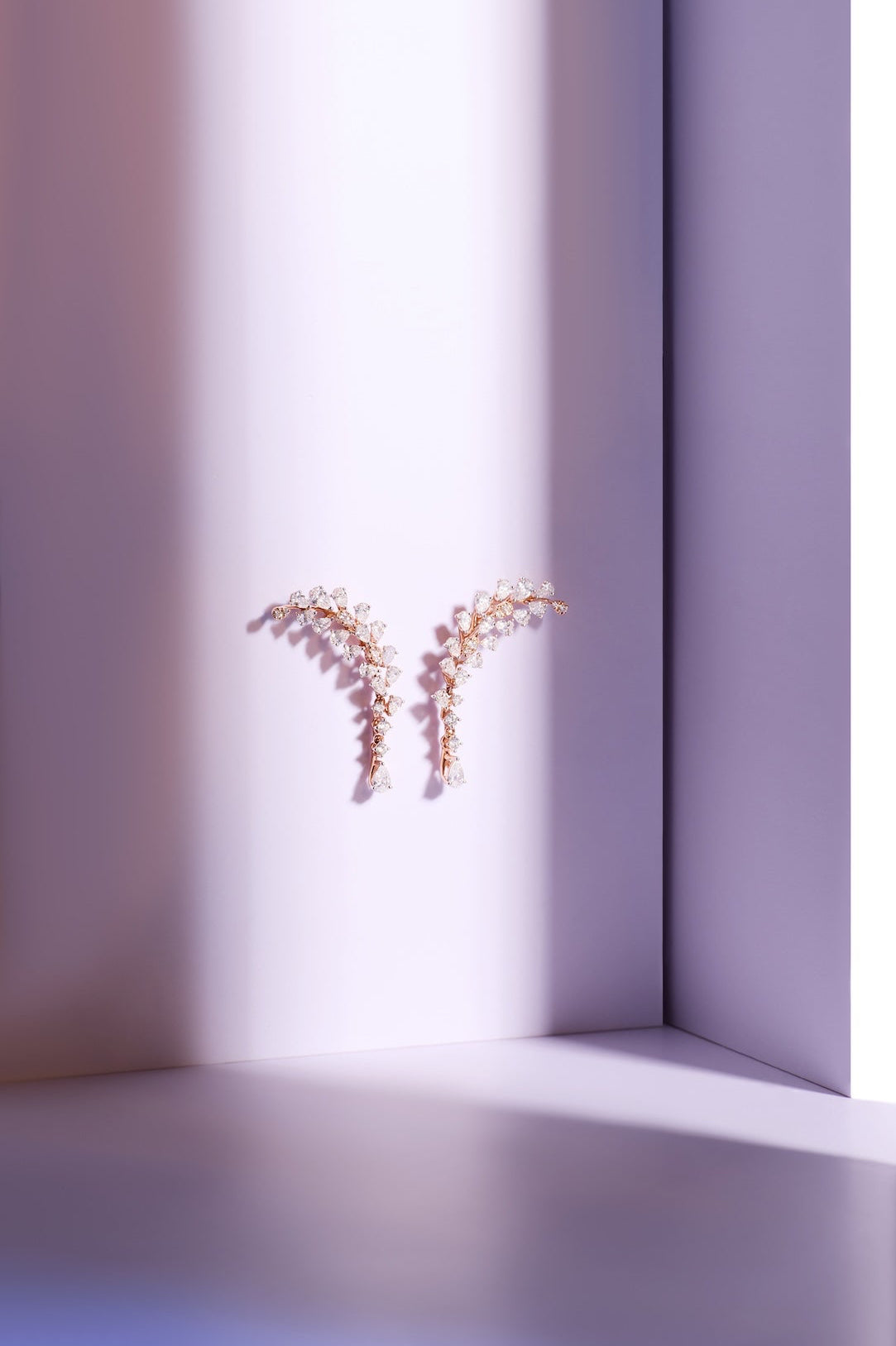 A pair of delicate gold-toned earrings with small clear stones arranged in a Y-shape, displayed on a minimalist lavender and white studio set with dramatic side lighting.