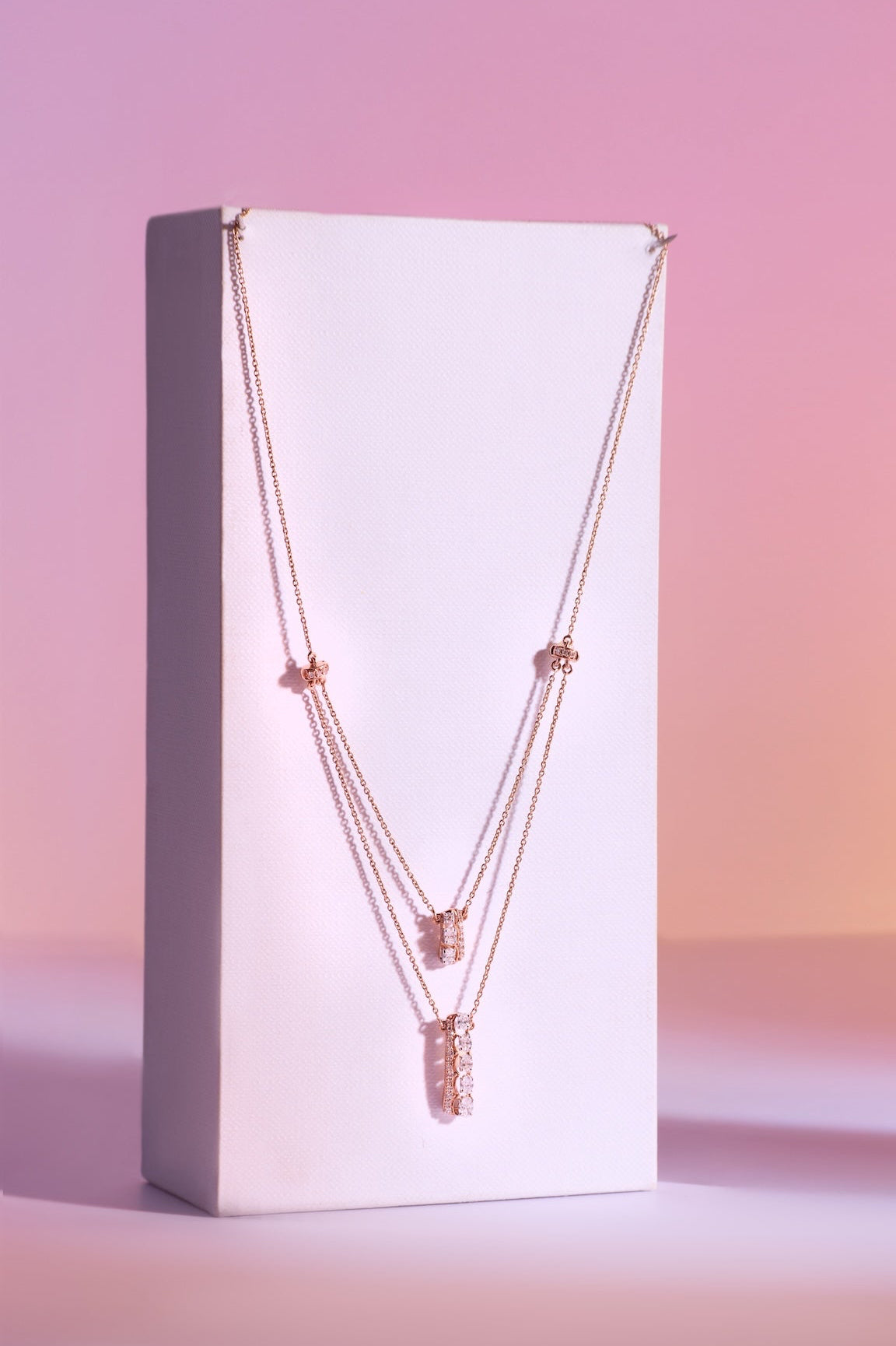 A rose gold multi-layer necklace with a textured pendant, displayed on a white rectangular stand against a pink gradient background.